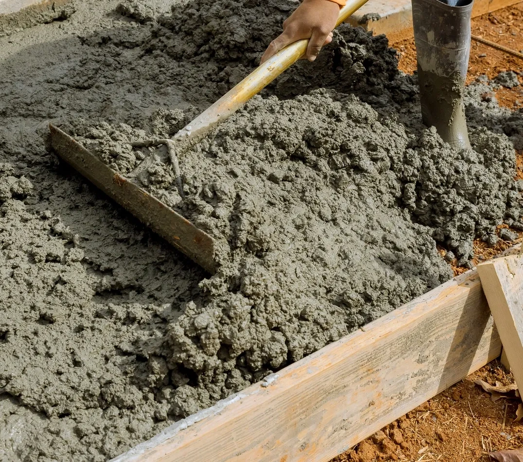 Person pouring concrete into a wooden box with a shovel, surrounded by mud and wooden pieces