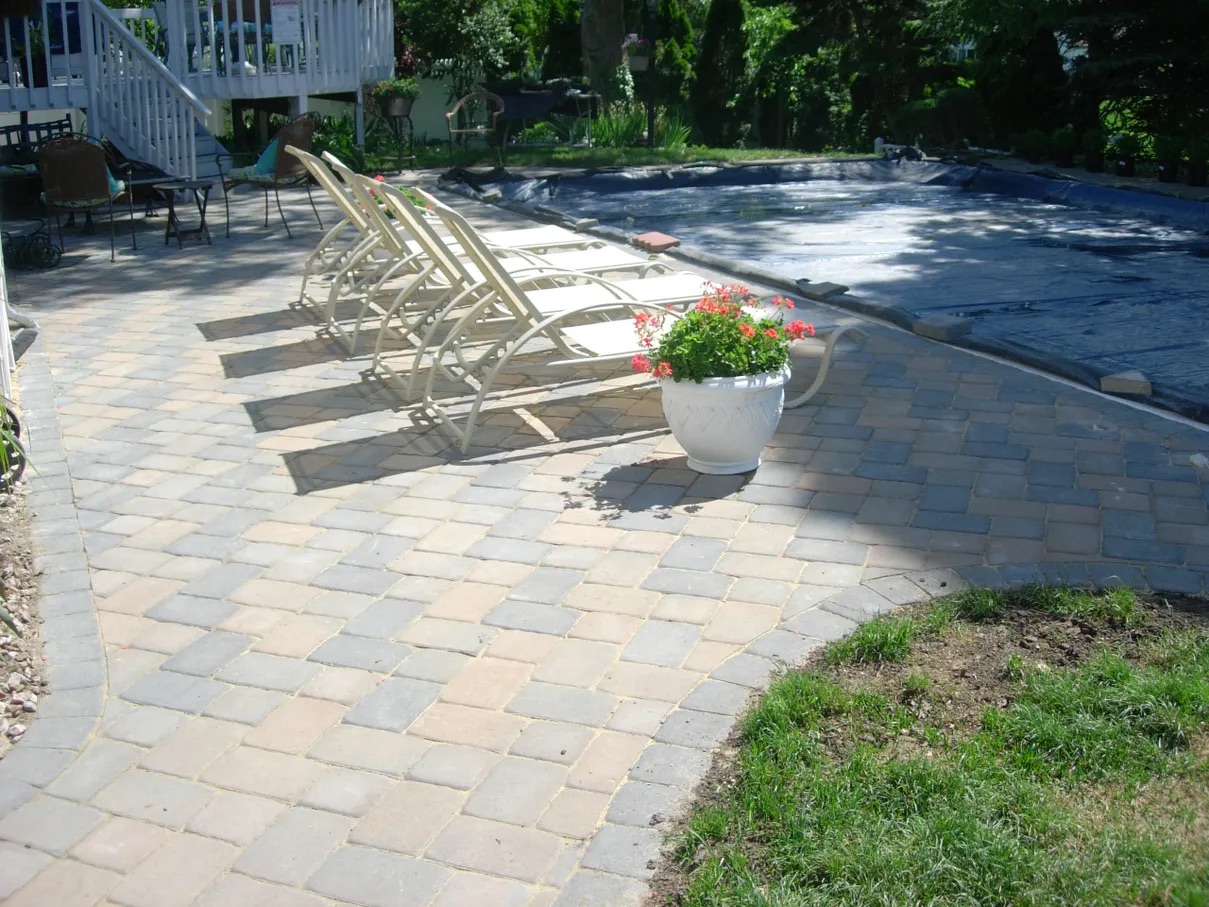 Brick patio with chairs and a pool in the background, surrounded by lush green grass and trees