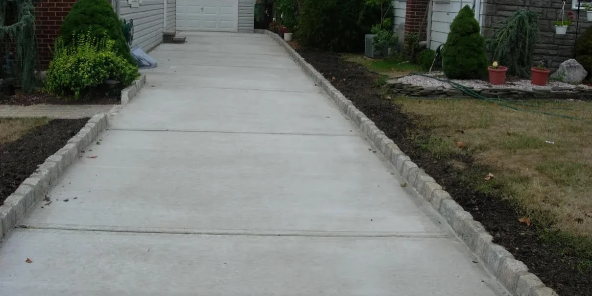 Concrete driveway with a curb in front of a house, surrounded by buildings with windows, trees, plants, grass, and pots