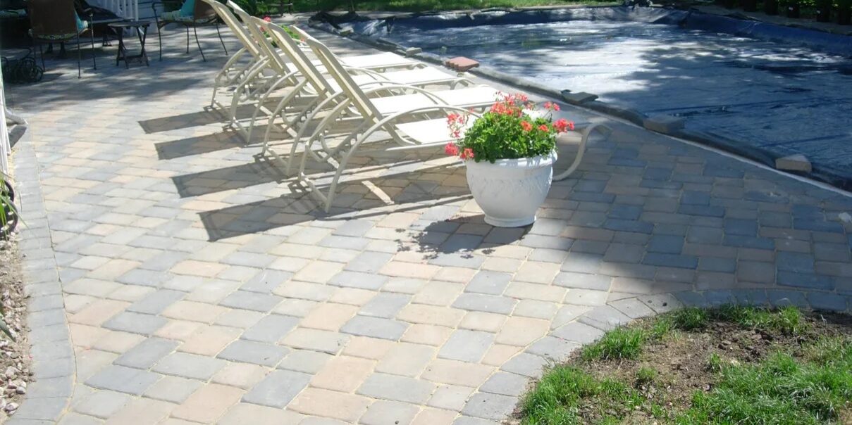 Brick patio with chairs and a pool in the background, surrounded by lush green grass and trees